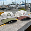 Photo image (left to right) of cream with olive logo and cream with brown dad hats with contrasting brims and embroidered logo outside on picnic table.