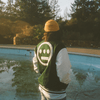 A side view of a man standing beside an abandoned swimming pool, wearing a green varsity jacket with white sleeves and a large white Hieroglyphics hip-hop logo on the center of the back.
