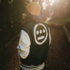 A man standing outdoors, back to the camera, wearing a green varsity jacket with white sleeves and a large white Hieroglyphics hip-hop logo on the center of the back.