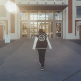 A man walking outdoors, back to the camera, with a green varsity jacket with white sleeves and a large white Hieroglyphics hip-hop logo on the center of the back thrown over his shoulders.